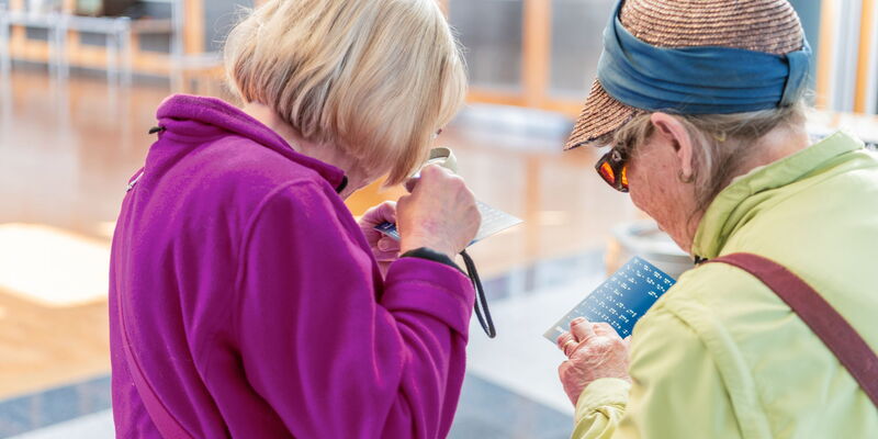 Zwei ältere Damen sehen sich Informationskarten an – eine benutzt eine Lupe und eine liest Braille-Schrift